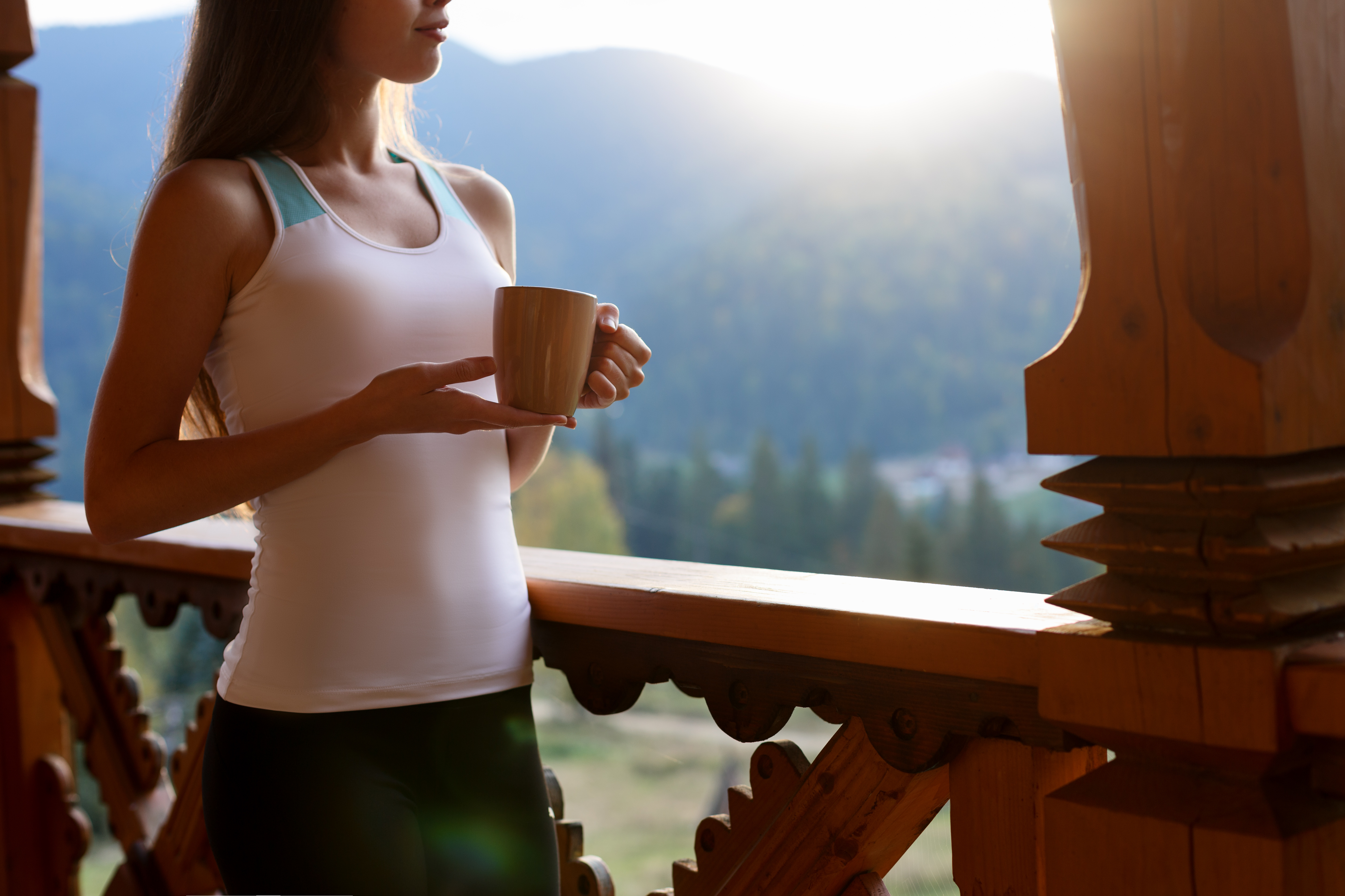 Slim caucasian woman holds cup of tea in her hands at mountain resort. Sports girl with hot coffee mug at wooden balcony of country house. Forest and mountains on background Dietary concept.